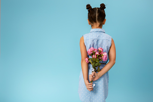 little child girl holding bouquet of roses behind her back on blue background. back view. copy space