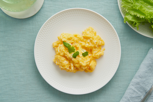 Scrambled eggs, omelette. Breakfast with pan-fried eggs. Texture of omelet on white plate on green mint linen textile tablecloth. Keto ketogenic diet. Soft light, top view
