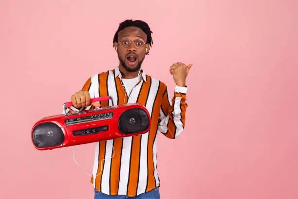 Shocked surprised african man with dreadlocks in striped shirt holding tape recorder listening to music in earphones pointing at freespace for advertisement. Studio shot isolated on pink background