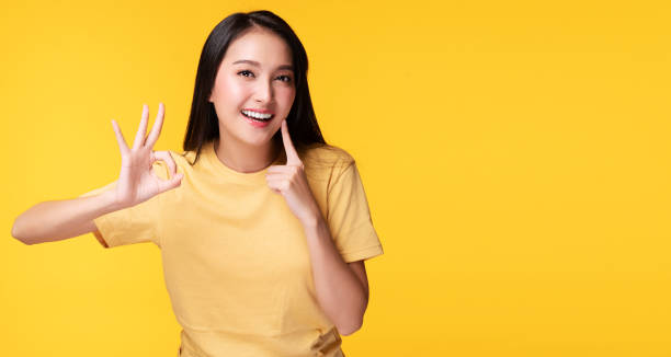 istock Cheerful young woman using finger point to her whitening teeth while make hand sign ok over isolated yellow background. She showing her cleaning fresh toothy. Dental care and healthy concept. 1303832916