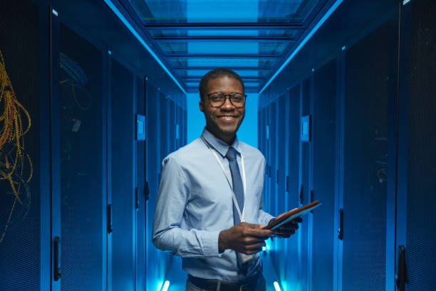 Smiling African American Man in Data Center Waist up portrait of smiling African American man standing by server cabinet while working with supercomputer in data center and holding tablet, copy space blue business stock pictures, royalty-free photos & images