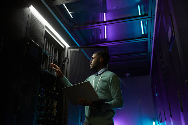 African American IT Engineer in Data Center Low angle portrait of young African American data engineer working with supercomputer in server room lit by blue light and holding laptop, copy space security system photos stock pictures, royalty-free photos & images