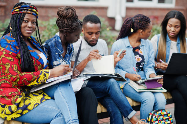 Group of five african college students spending time together on campus at university yard. Black afro friends studying at bench with school items, laptops notebooks.