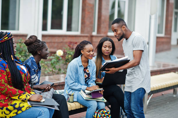 Group of five african college students spending time together on campus at university yard. Black afro friends studying at bench with school items, laptops notebooks.