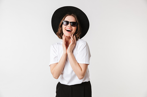 Portrait of a laughing young woman dressed in black hat and sunglasses posing while standing isolated over white background