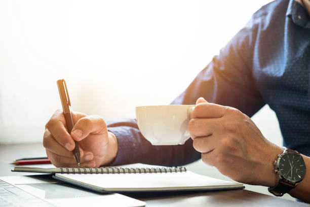 istock man hand holding coffee cup with writing pen memo into notebook paper on table desk office. Workplace for business and student, writer with copy space. business working and learning education concept. 1303844098