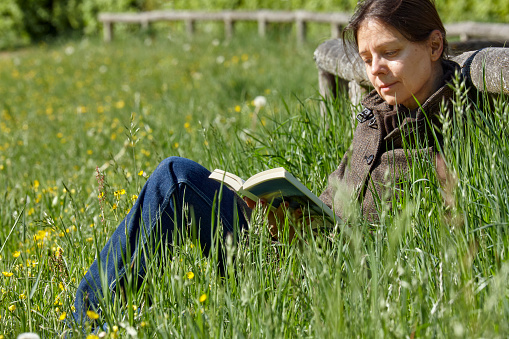 Full frame daylight image of a mature woman reading a book while sitting in a meadow in front of a transverse wooden tree trunk as a fence