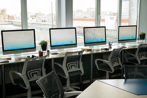Row of computer monitors standing on desks against large windows inside contemporary empty call center at the end of working day