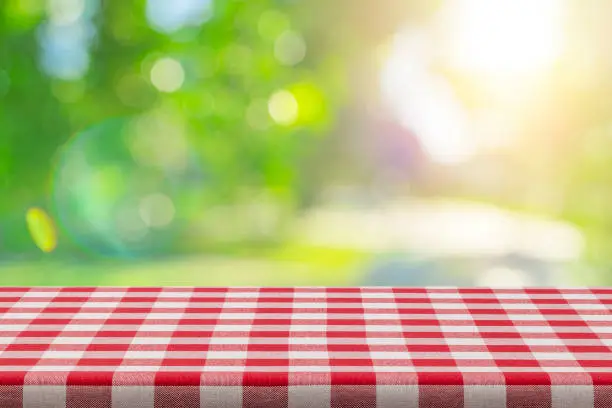 Spring backgrounds: empty table covered with red and white picnic tablecloth and defocused lush foliage at background. Ideal for product display on top of the table. Predominant color are green, yellow and red.