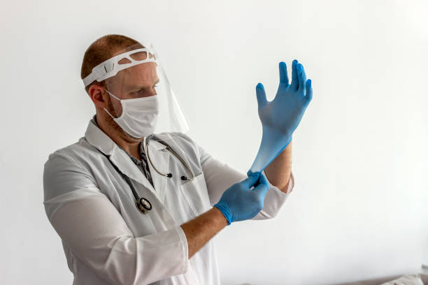 Caucasian young doctor putting surgical gloves on over white background. Caucasian young doctor or male nurse in white uniform and face protective medical mask putting surgical gloves on over white background. Healthcare, profession and medicine concept. Copy space. putting on latex gloves stock pictures, royalty-free photos & images