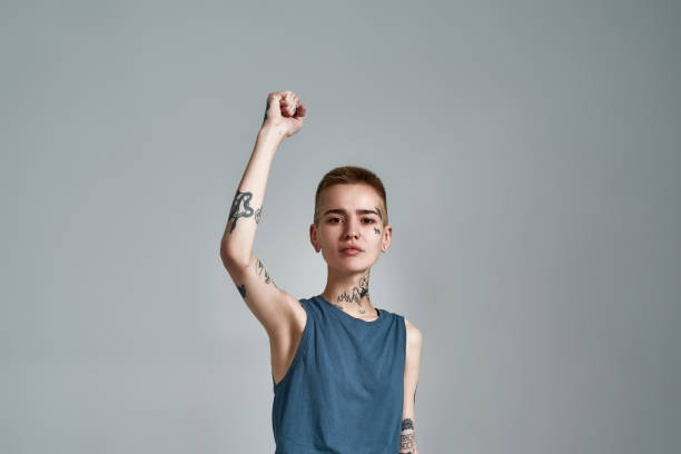 istock Cute tattooed young caucasian woman protester with piercing looking at camera while posing with raised arm isolated over gray background 1303871541