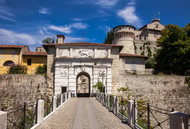 Main entrance to Castle of Brescia (Castello di Brescia) or Falcon of Italy (Falcone d'Italia) in Brescia, Lombardy, Northern Italy. Footbridge over the moat leads to the white marble arched gate, decorated with Venetian winged lion