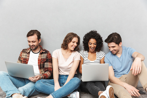 Photo of happy group of friends sitting isolated over grey wall background using laptop computers.