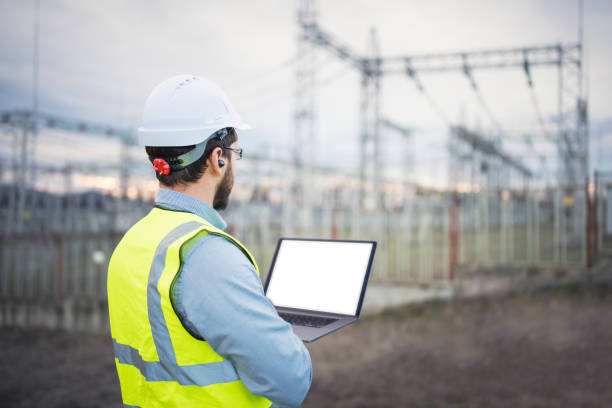Portrait of confident male engineer using a laptop in front of electric power station. Rear view of an electrical engineer using a laptop and hands-free device while working. electricity substation photos stock pictures, royalty-free photos & images