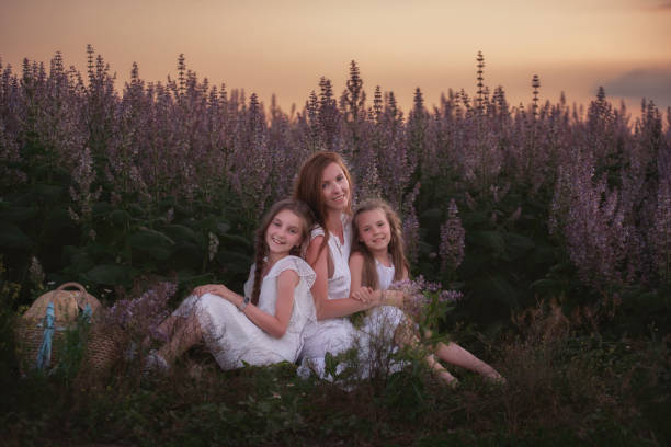 Young mother embraces her two daughters among a purple blooming sage field. Dressed in white with straw hats. Woman lovingly hugs girls, strokes, kisses. Out-of-town walks, weekend travel. Copy space la joven madre abraza a sus dos hijas entre un campo de salvia púrpura floreciente. vestido de blanco con sombreros de paja. mujer abraza con amor a chicas, golpes, besos. paseos fuera de la ciudad, viajes de fin de semana. copiar espacio