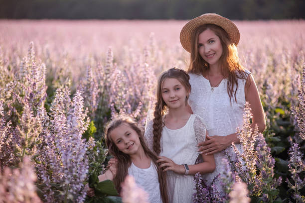 istock Young mother embraces her two daughters among a purple blooming sage field. Dressed in white with straw hats. Woman lovingly hugs girls, strokes, kisses. Out-of-town walks, weekend travel. Copy space 1303893473