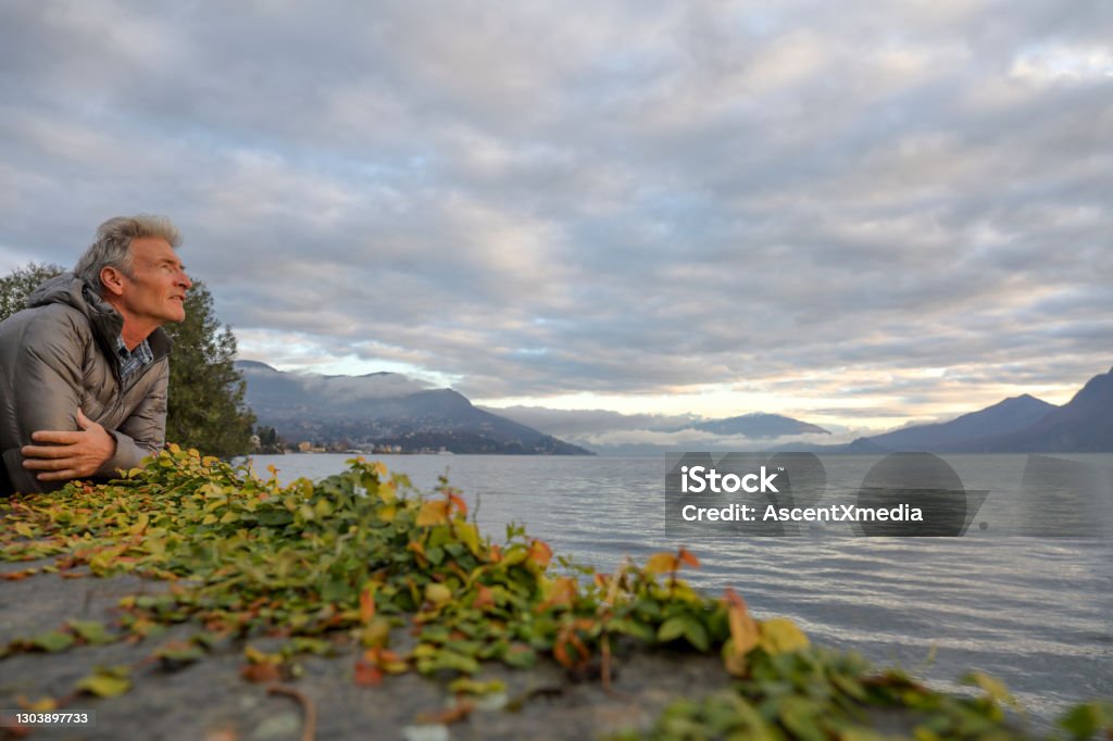 Mature man watches sunrise over lake and mountains He leans on a balcony Autumn Stock Photo