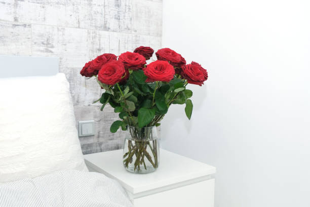 istock Beautiful red rose bouquet flowers on white bed-side table by bed with white pillow on background of gray grunge wall. Light minimal interior, decoration of bedroom, copy space 1303899032