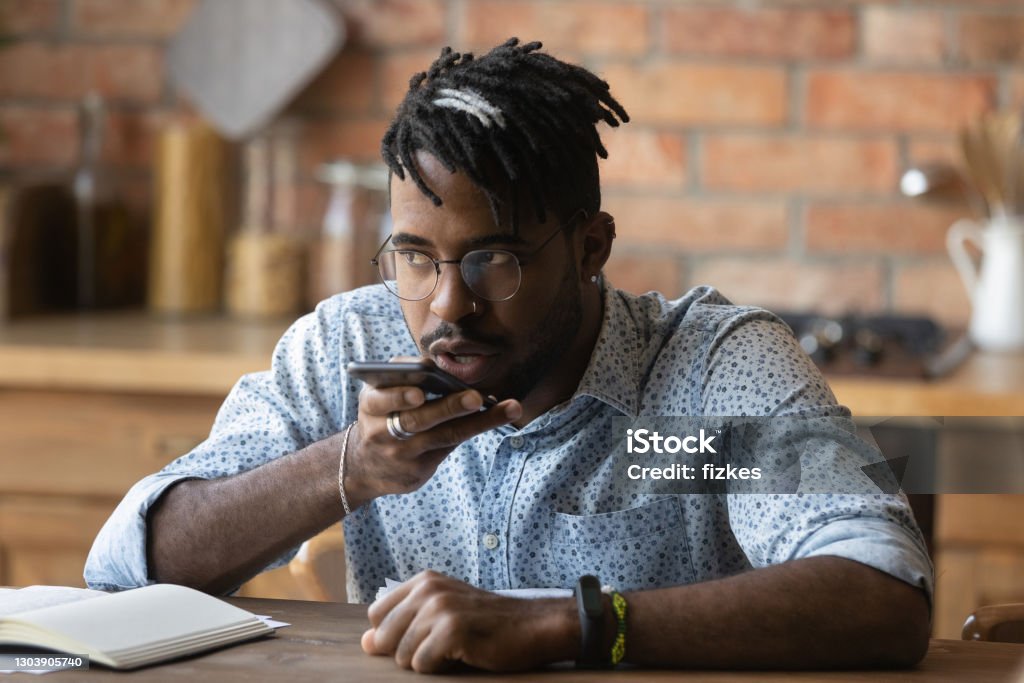 Close up serious African American man recording voice message Close up serious African American man wearing glasses recording voice message, holding phone near mouth, speaking, activating digital assistant, searching in internet, sitting at table at home Telephone Stock Photo