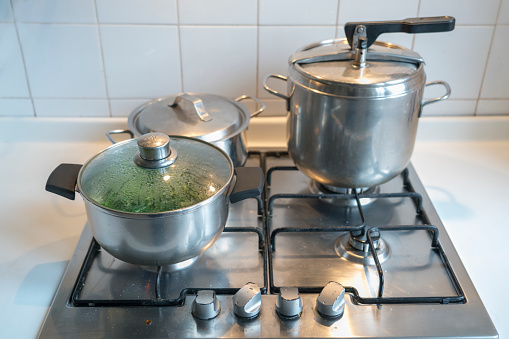 Cooking vegetable and other food on gas stove in home kitchen. Large pressure cooker and smaller pot, utensils.