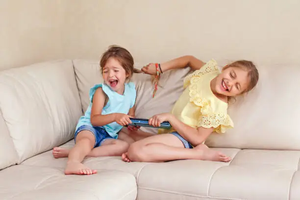 Photo of Two little mad angry girls sisters having fight at home. Friends girls can not share a gadget tablet. Lifestyle authentic funny family moment of siblings quarrel life. Kids bad behaviour.
