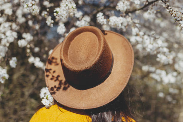 istock Stylish woman in hat posing at blooming cherry branches in sunny spring, back view. Tranquil moment 1303909809