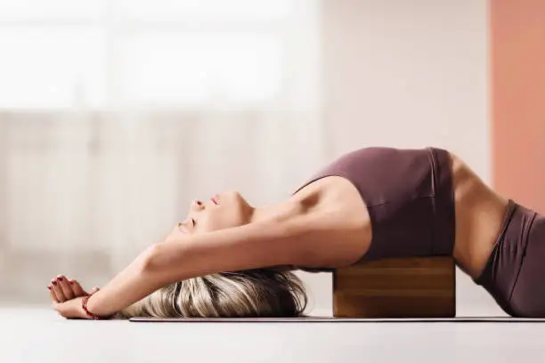 Beautiful blonde woman in sportswear, lies on a wooden block for yoga to improve posture.