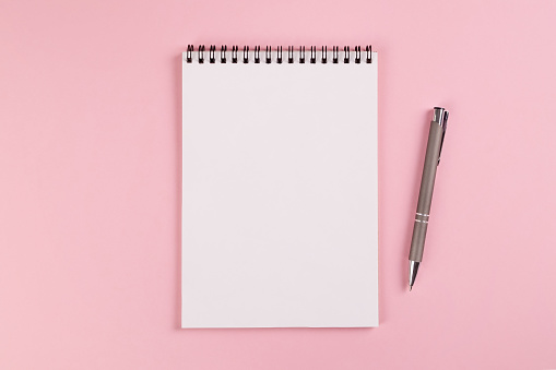 A notebook with a blank page and a ballpoint pen lie on a pink background. Copy space, layout, mockup.