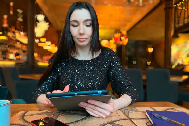 une fille aux cheveux bruns travaille dans un café avec une tablette, un téléphone et un ordinateur portable sont à proximité. coworking, organisation du flux de travail. concept d’affaires. - women computer home interior brown hair photos et images de collection