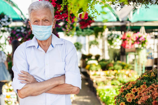 istock Senior man sells flowers on the market stall. He is wearing pulled down protective face mask 1303926418