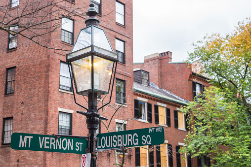 Close up of a traditional gas lit lamp post with street signs at a crossroads in a historic residential district. Traditional brick houses are visible in background. Beacon Hill, Boston, MA, USA.
