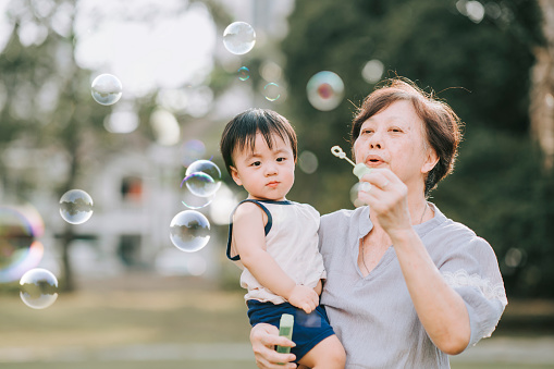 asian chinese grandmother playing bubble together with her grandson in public park during weekend evening backlit