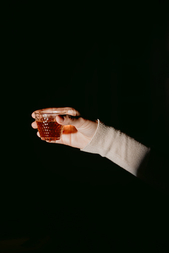 A close-up shot of an unrecognizable woman's hand, holding a small glass cup of tea against a black background.