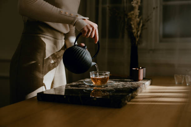 Anonymous Woman Enjoying a Slow Afternoon, Carefully Pouring herself Tea An unrecognizable woman slowly pouring tea from a black teapot into a glass cup. steep stock pictures, royalty-free photos & images