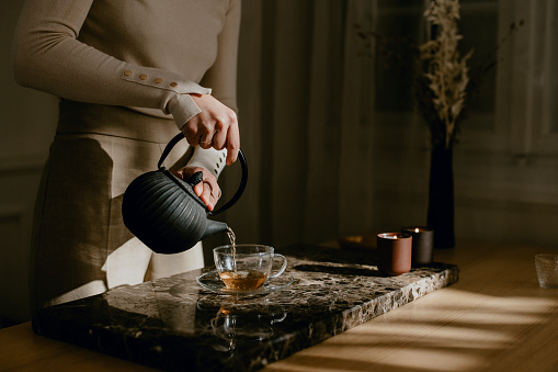 An unrecognizable woman slowly pouring tea from a black teapot into a glass cup.