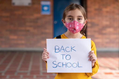 Portrait of a girl wearing a facemask at school during the COVID-19 pandemic and holding a back-to-school sign - new lifestyle concepts