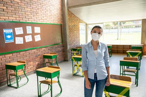 Latin American teacher in a school classroom wearing a facemask during the COVID-19 pandemic and following guidelines of social distancing. **DESIGN ON POSTER WAS MADE FROM SCRATCH BY US**