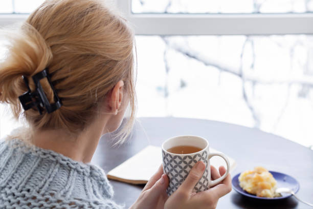 una donna con una tazza di tè è seduta vicino alla finestra - snow heat adult blond hair foto e immagini stock