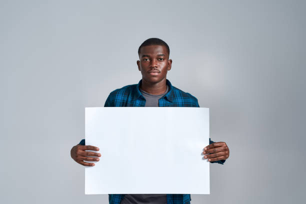 istock Serious young african american guy in casual clothes looking at camera, displaying blank banner ad, holding it in front of him, posing isolated over gray background 1303971443