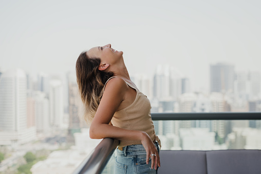 Happy young woman in denim shorts and beige crop top standing at the balcony. Positive attitude. Happy and smiling face. Relaxation and enjoyment concept. Stress free