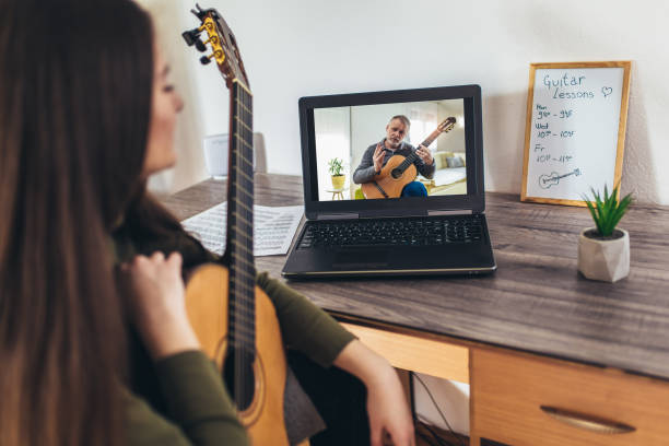 istock Focused girl playing acoustic guitar and watching online course on laptop while practicing at home. 1303975314