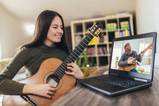 istock Focused girl playing acoustic guitar and watching online course on laptop while practicing at home. 1303975336