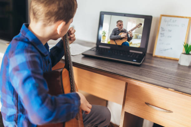 istock Focused boy playing acoustic guitar and watching online course on laptop while practicing at home. 1303976290