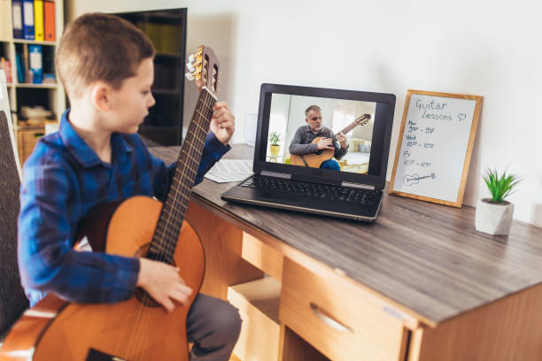 istock Focused boy playing acoustic guitar and watching online course on laptop while practicing at home. 1303976377