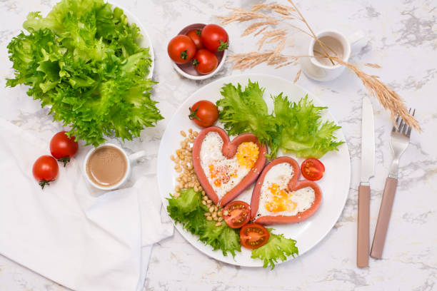 istock Romantic breakfast. Fried eggs in heart shaped sausages, lettuce and cherry tomatoes on a plate on the table. Top view 1303985668