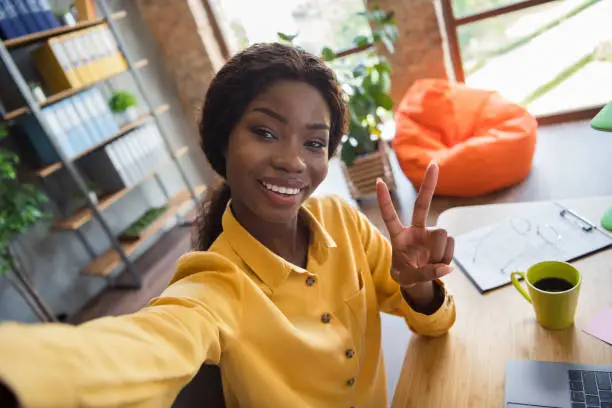 Photo portrait of african american girl taking selfie showing v-sign at workdesk in modern office indoors.