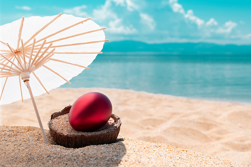 Red Easter egg on coconut shell on golden sandy beach under sun umbrella and blurred blue sea and sky in background. Celebrating Easter holiday on tropical countries beach Greeting card, copy space