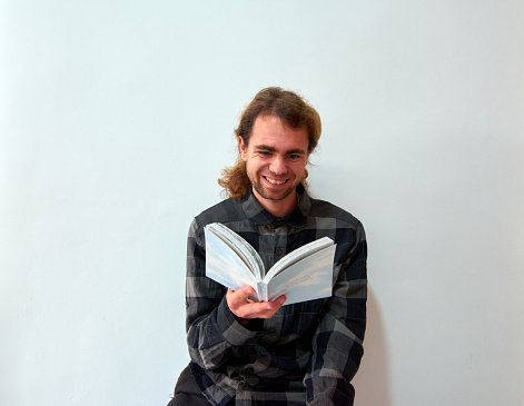 A handsome Caucasian man with long hair happily reading a book on the background of the white wall