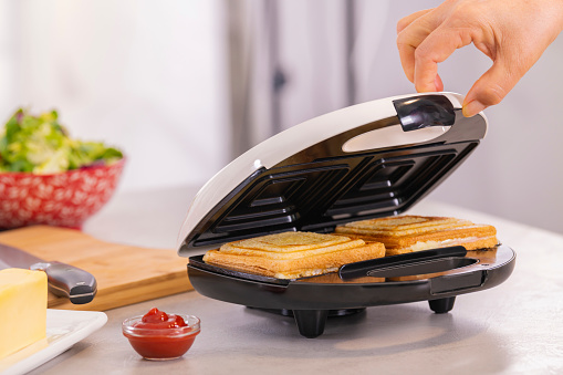 Female hand carefully opening the lid of a sandwich making machine with hot sandwiches. Selective focus and close up. Homemade food concept.