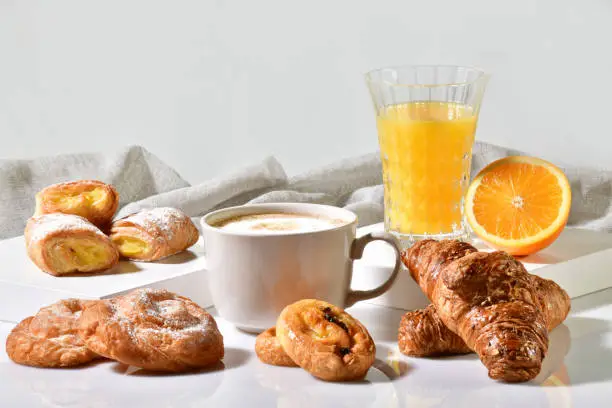 Photo of Cup of coffee surrounded by delicious pastries, with an orange juice and half orange at the back on a light background. Traditional breakfast concept.
